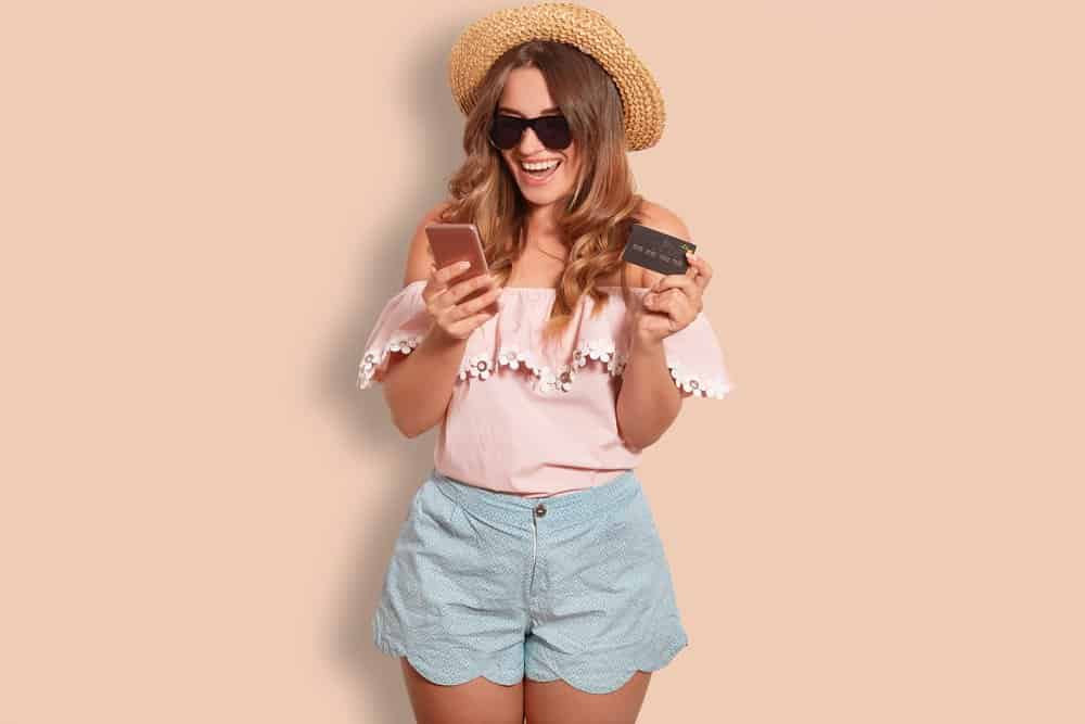 Glad young lady makes purchases online, uses modern application, holds cell phone, wears trendy sunglasses, fashionable clothes, poses against studio background. People and online shopping concept